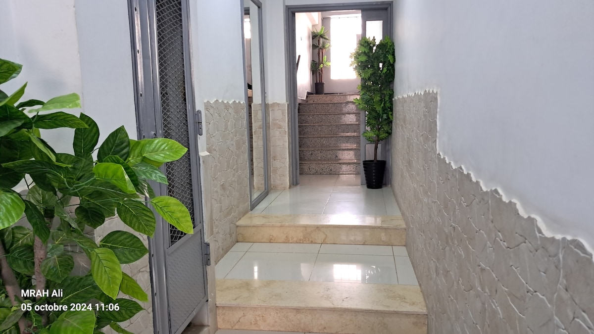 The hallway features polished tiles and a staircase leading to an upper level. Lush green plants in pots add a touch of nature, while glass doors provide an inviting entrance. Neutral wall colors enhance the open and well-lit ambiance.