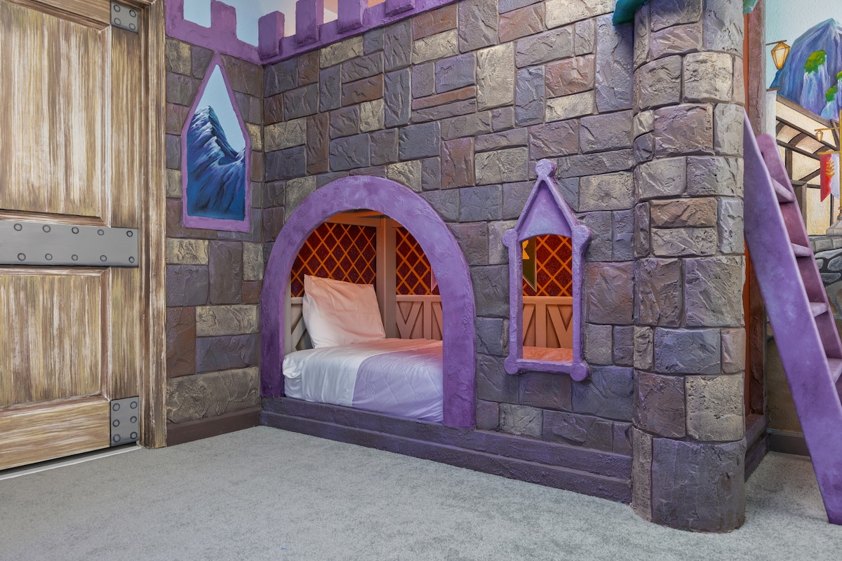 A themed bedroom features castle-like elements, with textured stone walls and colorful accents. A cozy sleeping area is highlighted under an archway with decorative windows, showcasing a spacious bed and inviting decor. A staircase leads to additional amenities in the room.