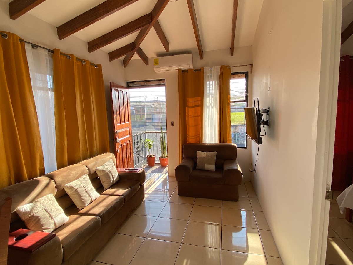 A spacious living area features a comfortable brown sofa and an armchair, positioned near large windows adorned with bright yellow curtains. Natural light fills the room, highlighting the tiled floor and wooden beam ceiling. A wall-mounted TV is visible, contributing to a cozy ambiance.