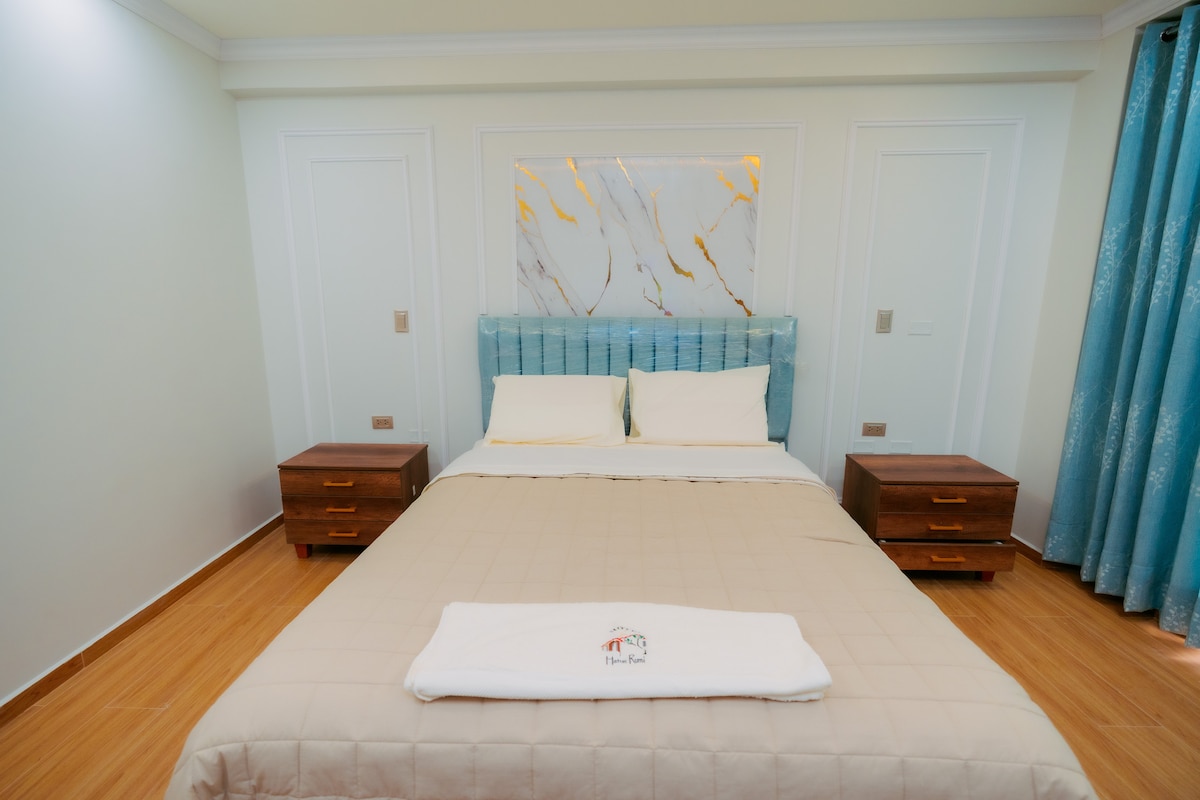 A well-appointed sleeping area features a spacious bed with neutral bedding. Two wooden nightstands flank each side, and a light-colored wall provides a subtle backdrop. Textured blue curtains frame the window, while a decorative wall piece adds a touch of elegance.