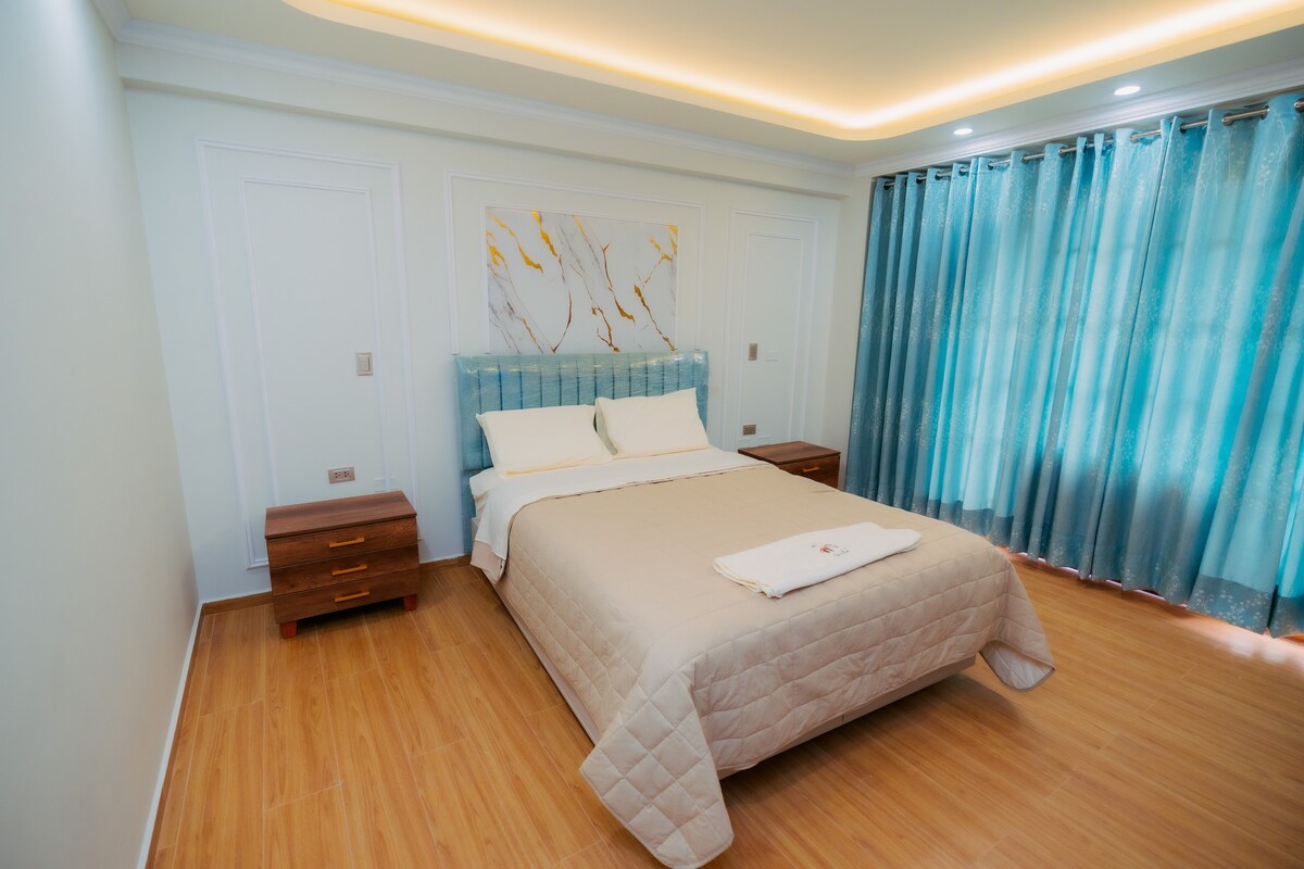 A well-lit bedroom features a spacious bed dressed in neutral linens. Flanking each side are wooden nightstands. The large windows are covered with sheer turquoise curtains, allowing natural light to fill the room, while a modern wall art piece adds a decorative touch.