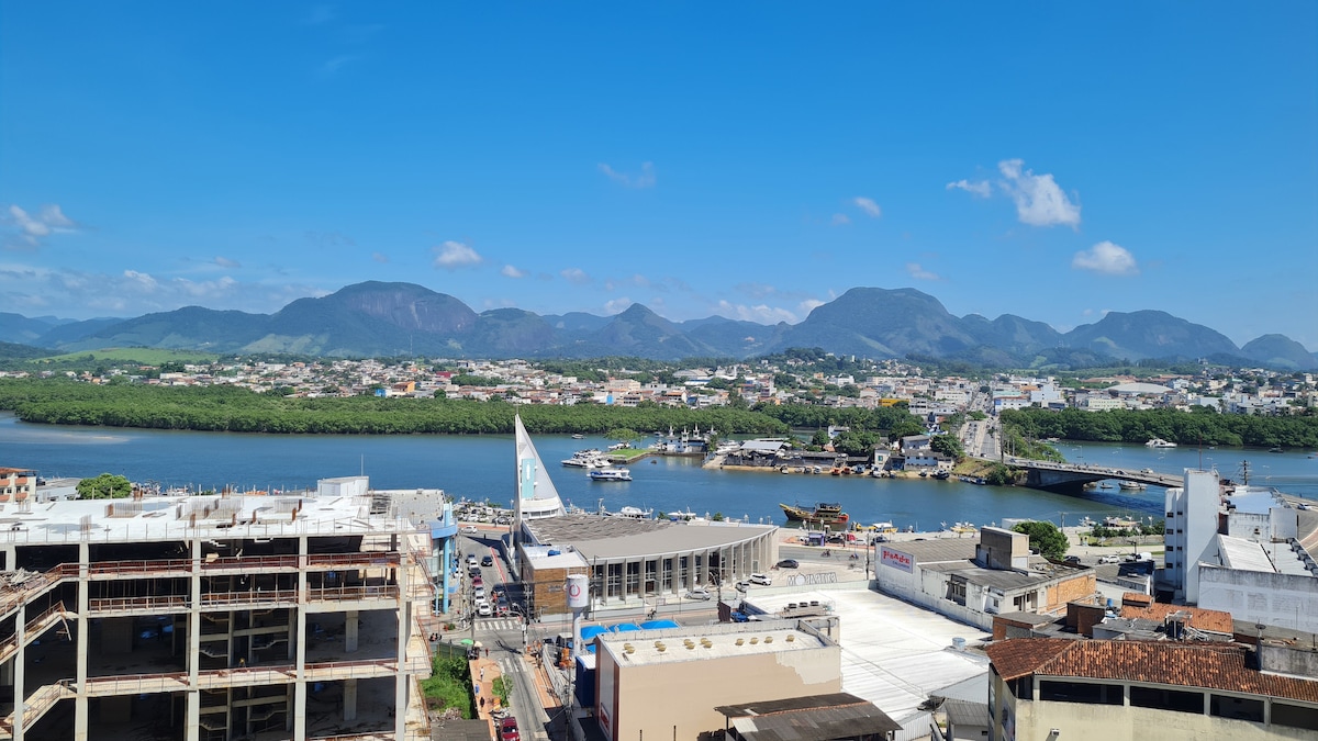 A panoramic view captures a vibrant city landscape bordered by lush mountains and blue skies. The river reflects the surroundings, while a mix of urban and natural elements occupies the scene, showcasing buildings and greenery along the water's edge.