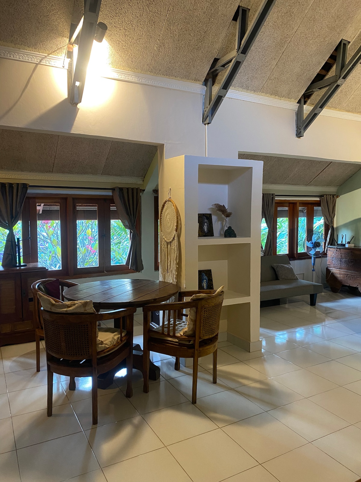 A dining area features a round wooden table surrounded by four woven chairs. Natural light filters through large windows adorned with soft curtains, illuminating the space. A decorative shelf stands nearby, displaying various ornamental items that add character to the room.