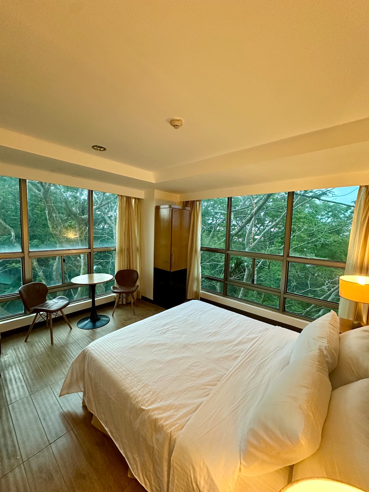 A bright bedroom features floor-to-ceiling windows offering views of surrounding trees. The spacious bed is dressed in crisp white linens. A small round table with two chairs is situated near the window, and a lamp beside the bed provides warm lighting.