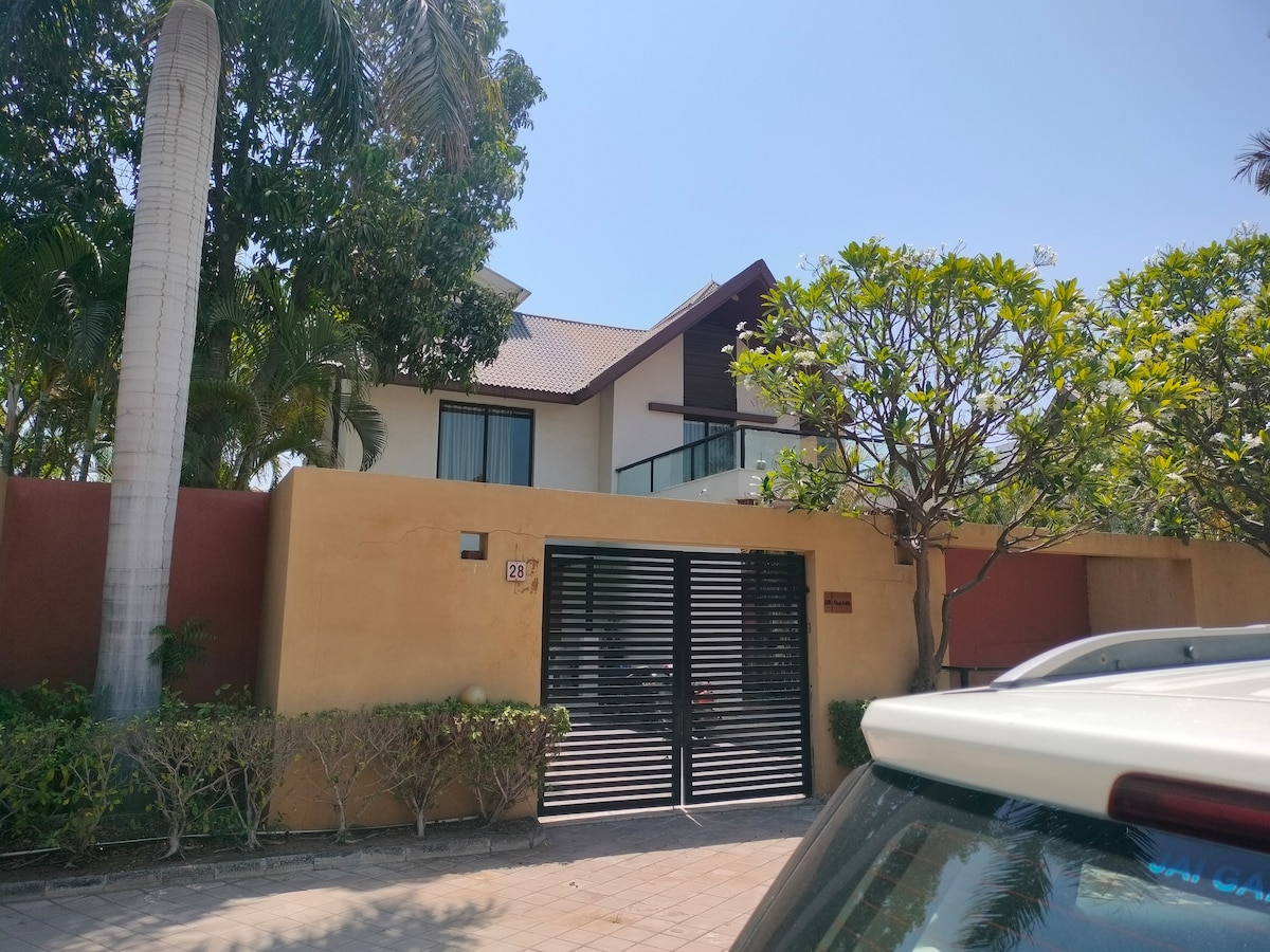 A modern bungalow is showcased behind a gated entrance, framed by lush greenery and palm trees. A bright blue sky is visible above, complementing the contemporary architecture with large windows and a sloped roof. The entrance is paved and lined with low shrubbery.