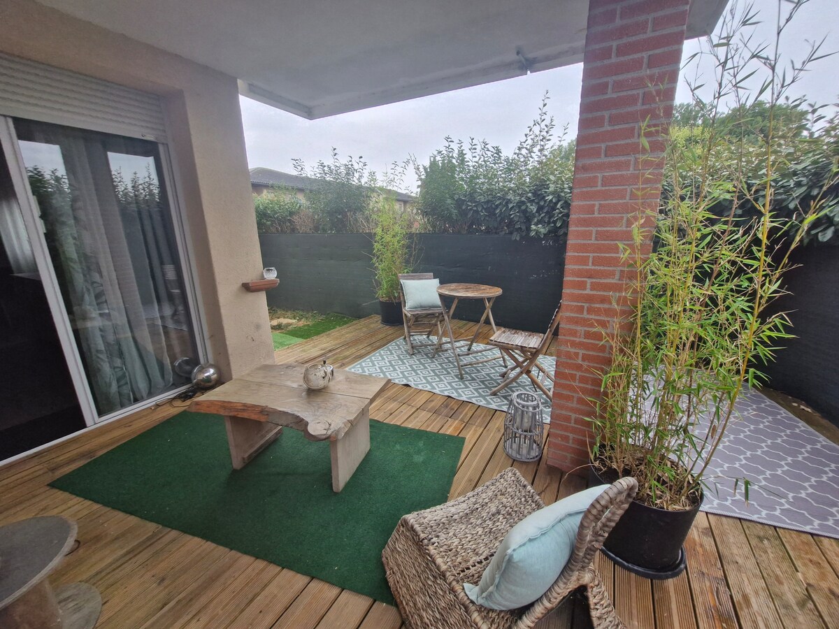 An outdoor terrace is shown, featuring a wooden table and two chairs arranged on wooden decking. Lush greenery borders the space, providing privacy. A small decorative lantern is positioned nearby, with a light rug complementing the natural elements.