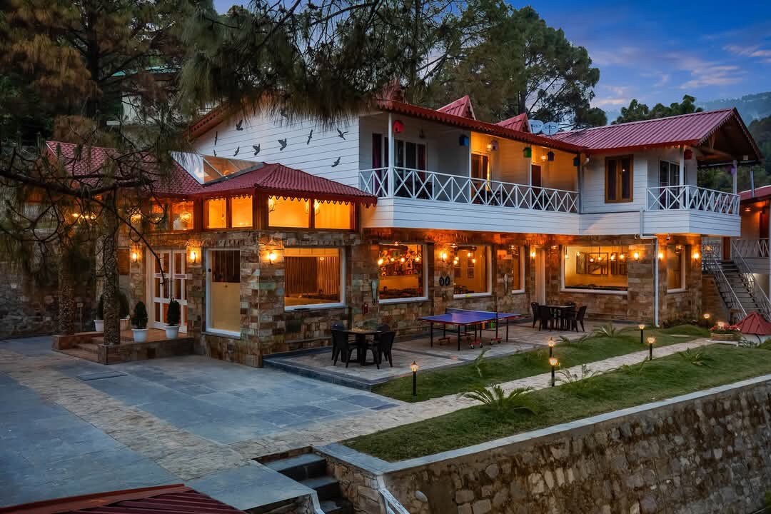 The exterior of a charming two-story structure is visible, featuring a combination of natural stone and wooden elements. A welcoming terrace is adorned with outdoor seating arrangements, complemented by warm lighting. Lush greenery surrounds the patio, creating a serene atmosphere amidst the hills.