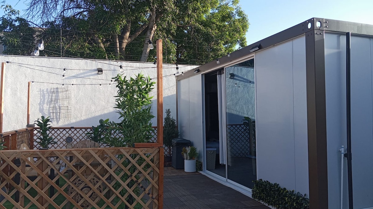 A modern container house is depicted with a wooden deck, surrounded by a lattice fence. Lush plants line the deck, and large glass doors reflect natural light from outside. String lights are visible overhead, contributing to a relaxed outdoor atmosphere.