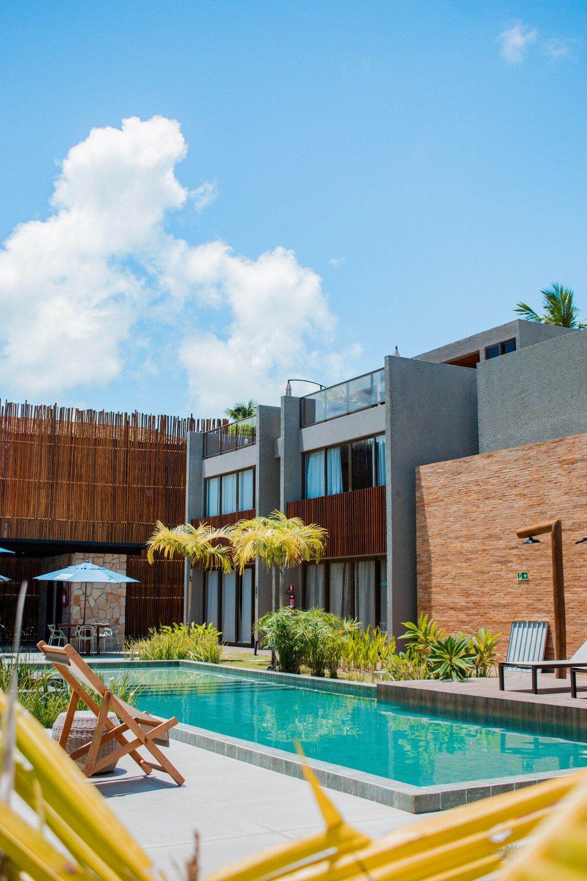 The exterior view of a modern building is showcased, featuring multiple units with large windows reflecting the sky. A tranquil pool is positioned in the foreground, surrounded by lush greenery, with sun loungers placed nearby for relaxation.