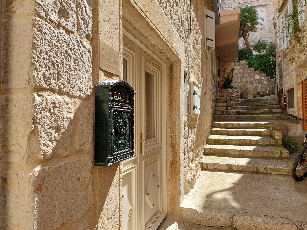 The entrance to the historic stone house features a wooden door with an ornate black mailbox mounted nearby. Natural stone walls create a rustic appeal, while stone steps ascend from the door. Lush greenery is visible in the background, enhancing the inviting pathway.