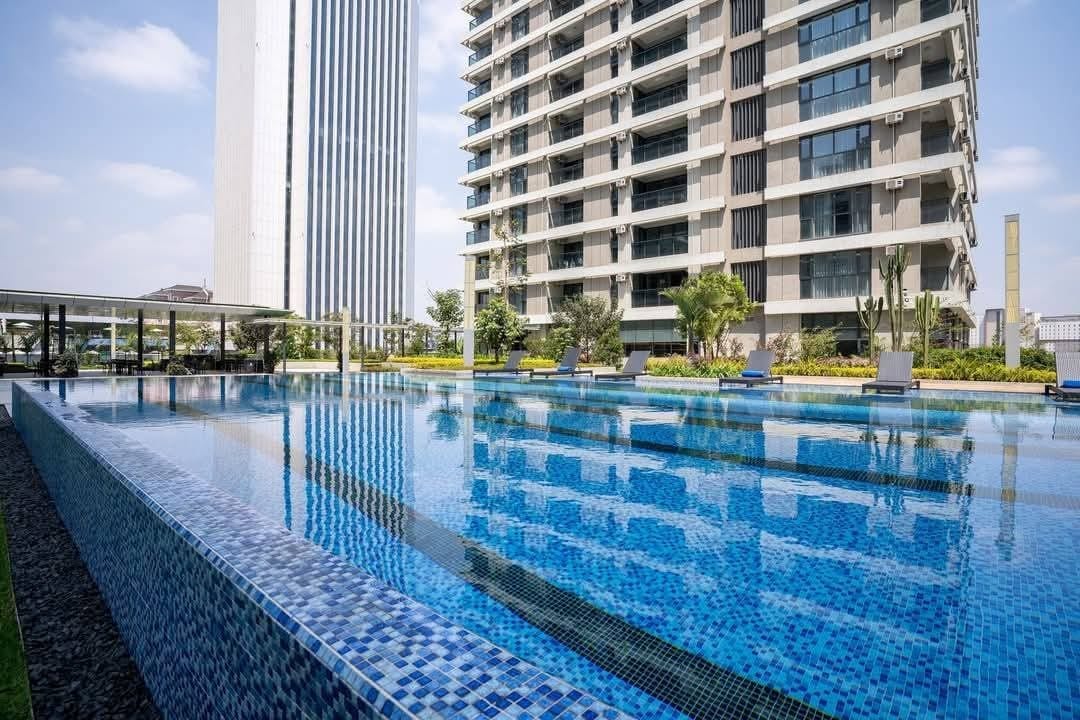 An outdoor swimming pool extends across a landscaped area, featuring various lounge chairs positioned along its edge. Tall buildings frame the scene, while vibrant greenery adds to the setting. The pool's surface reflects the clear blue sky above.