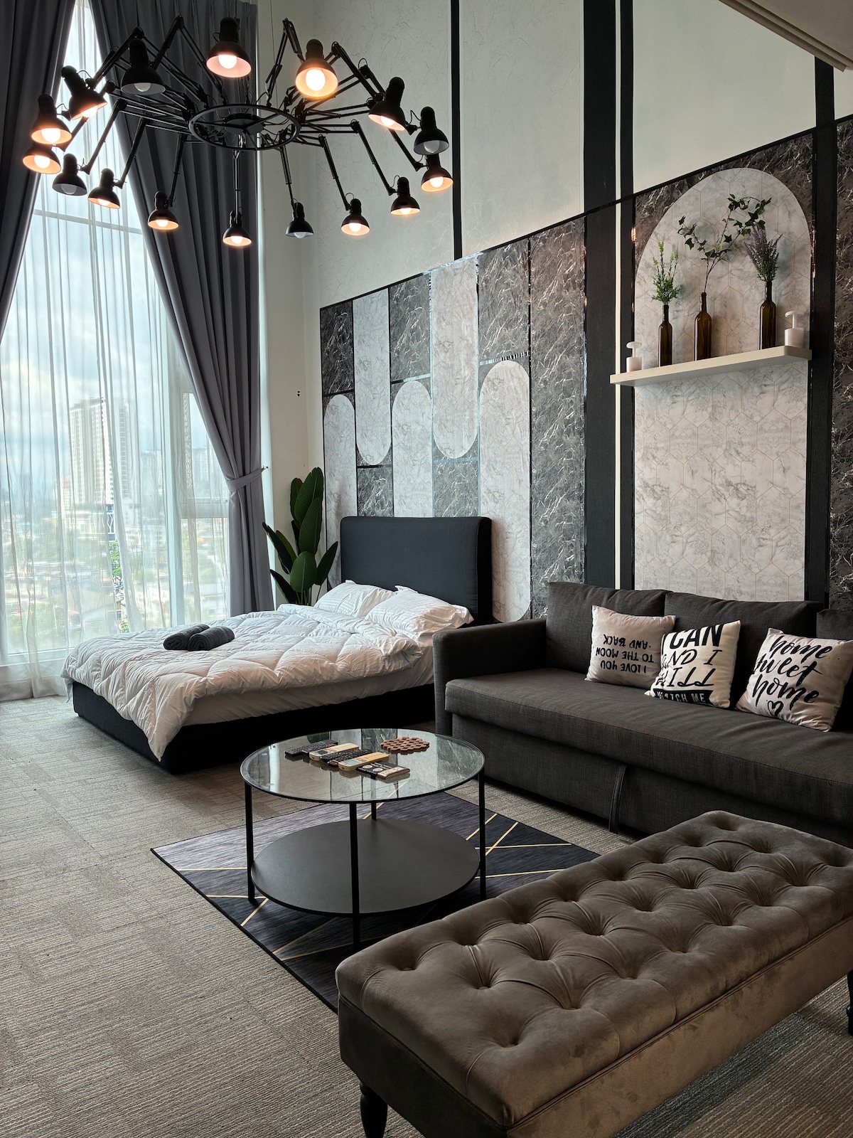 A stylish living area features a comfortable gray sofa and a tufted ottoman, complemented by a round coffee table. A spacious bed with white linens is positioned against a decorative wall, while large windows allow natural light to fill the room, framed by elegant curtains.