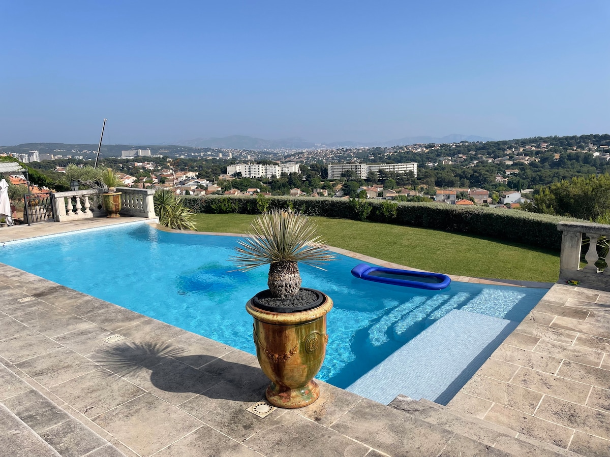 A refreshing salt-water pool is framed by a stone terrace, featuring a decorative pot with a plant. A blue inflatable float is visible in the water, while sweeping views of the surrounding hills and distant cityscape are enjoyed from the pool area.