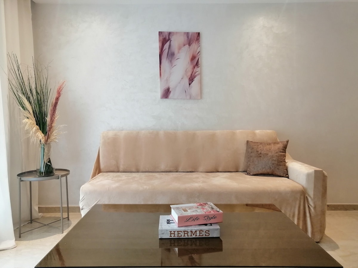 A light-colored sofa is positioned against a softly textured wall, complemented by a decorative cushion. A small round table holds stylish magazines. A minimalist wall hanging featuring pastel tones adds a subtle touch of elegance to the space, while a decorative plant provides natural interest.