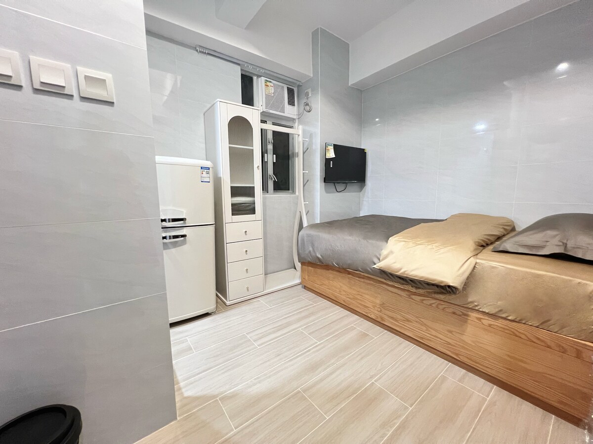 A modern studio features a spacious bed with neutral-toned bedding, accompanied by a minimalist wardrobe and a compact refrigerator. A wall-mounted television is positioned for viewing convenience. The sleek, tiled flooring adds to the contemporary appeal of the room.