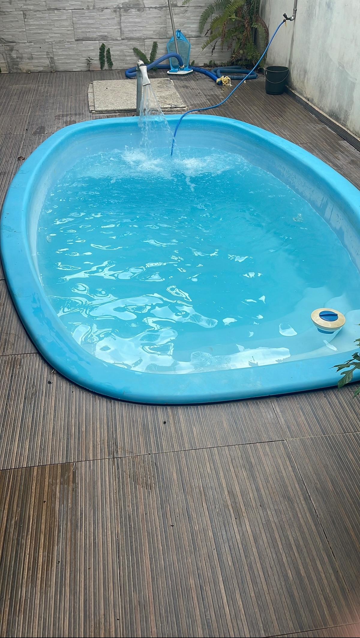 A serene swimming pool with clear blue water is surrounded by a wooden deck. Water gently flows from a small fountain, creating a calming sound. Lush plants and a light-colored wall contribute to a tranquil outdoor space.