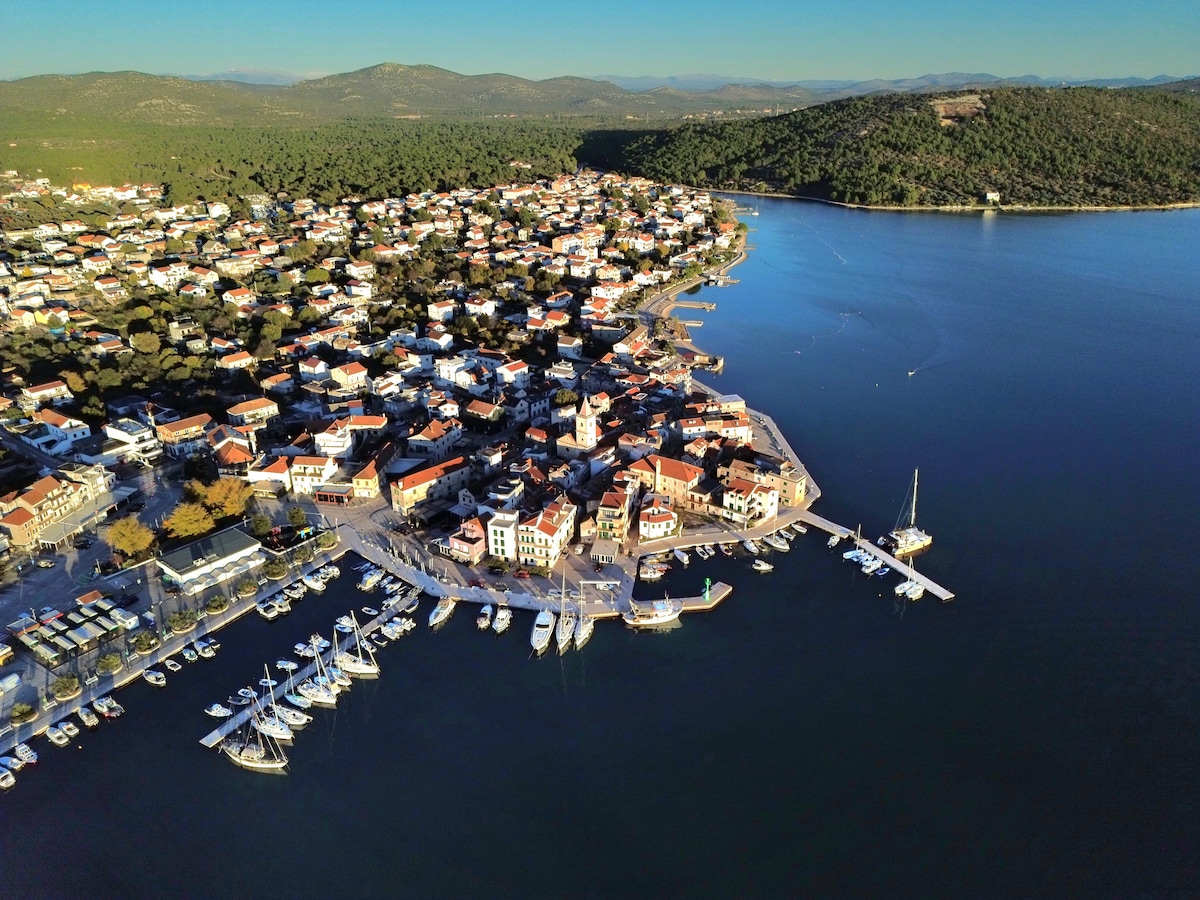 An aerial view of a coastal town features a picturesque waterfront area lined with boats and yachts. Colorful buildings with terracotta roofs curve along the shoreline, while green hills rise in the background. Clear blue water reflects the sky, enhancing the serene environment.