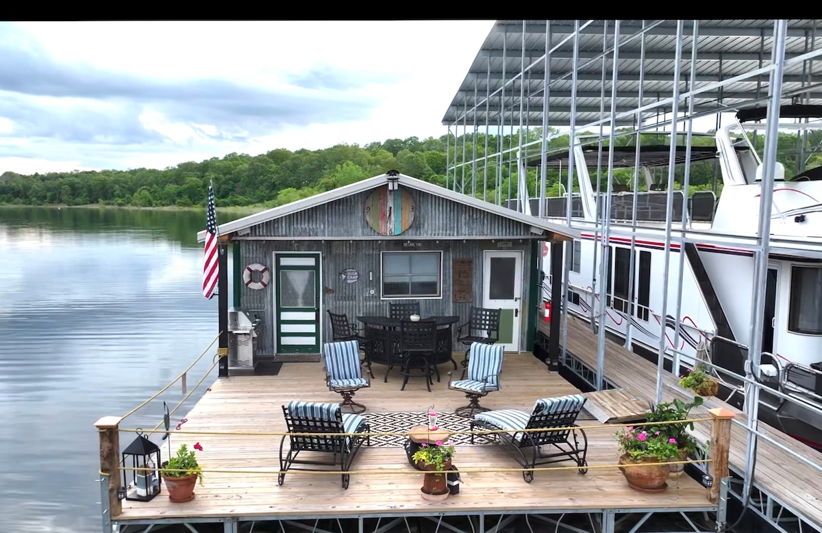 A spacious floating deck features lounge chairs and a dining area surrounded by potted plants. A flag is displayed on the side of the cozy cabin, with a view of the calm water and surrounding greenery. A boat slip is visible in the background.