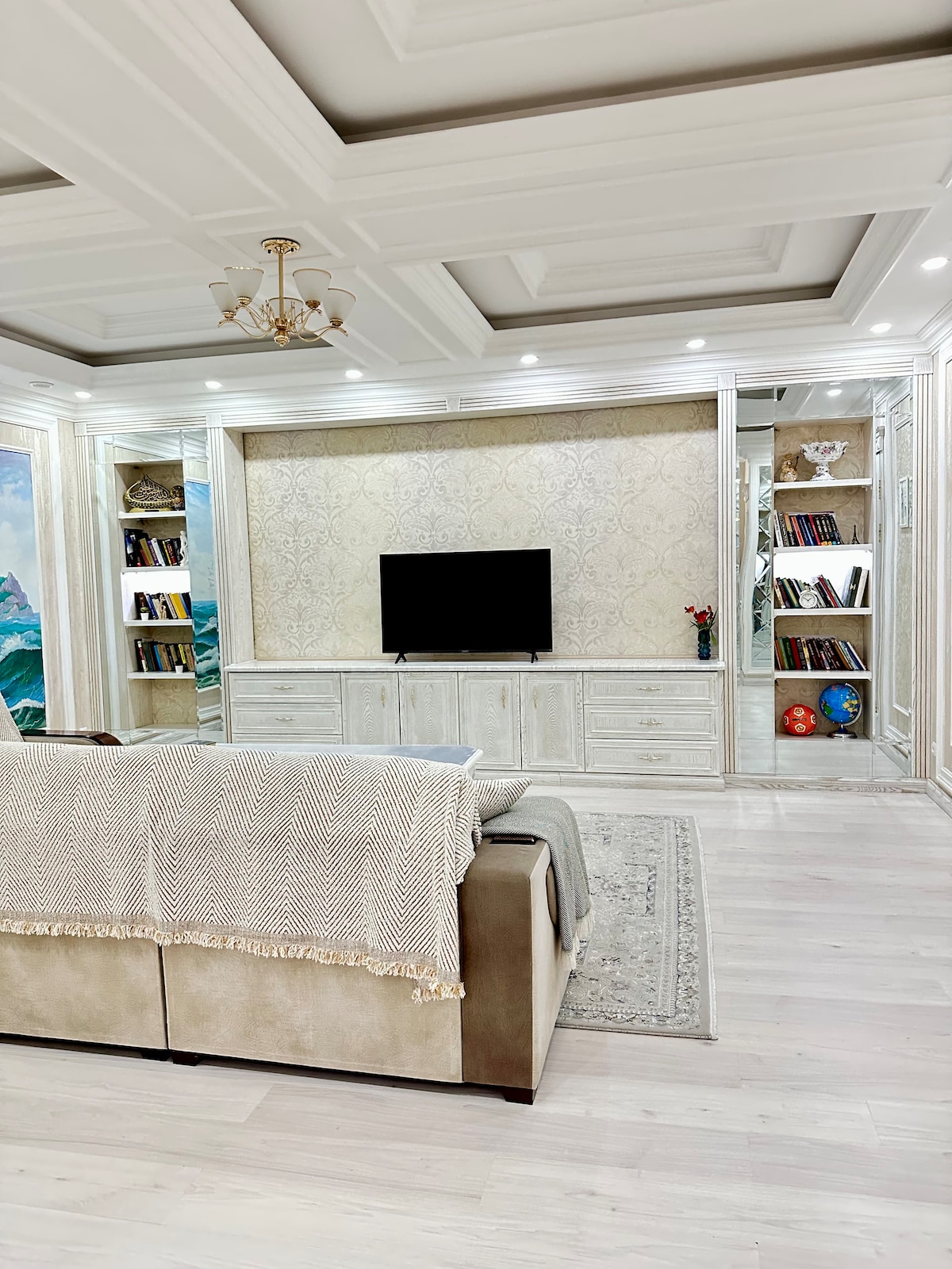 A spacious living area is presented with light-colored walls and an elegant coffered ceiling. A comfortable sofa is placed in front of a media console with a television. Bookshelves are built into the walls, showcasing a collection of books and decorative items.