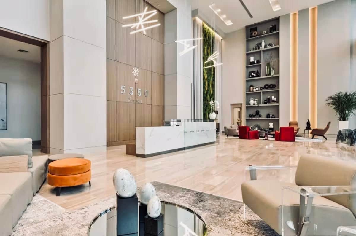 The modern lobby features a sleek reception desk made of light-colored stone, complemented by contemporary lighting fixtures. Comfortable seating areas include vibrant red chairs and a bright orange ottoman. Lush greenery is displayed on the walls, enhancing the inviting ambiance of the space.