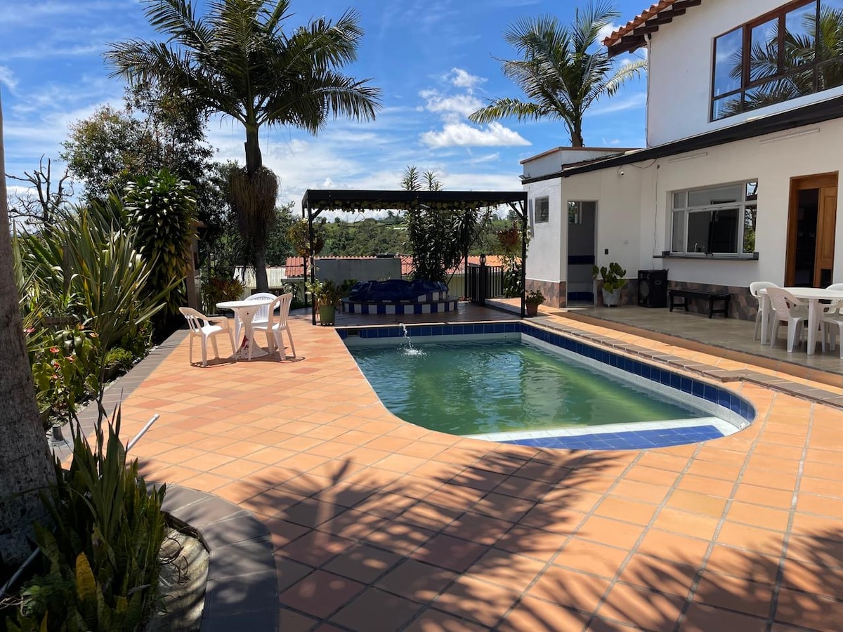 A private swimming pool is surrounded by tiled patio areas, with lounge chairs positioned under a pergola. Lush greenery, including palm trees, enhances the setting. A dining table with chairs is visible nearby, contributing to the inviting outdoor space.