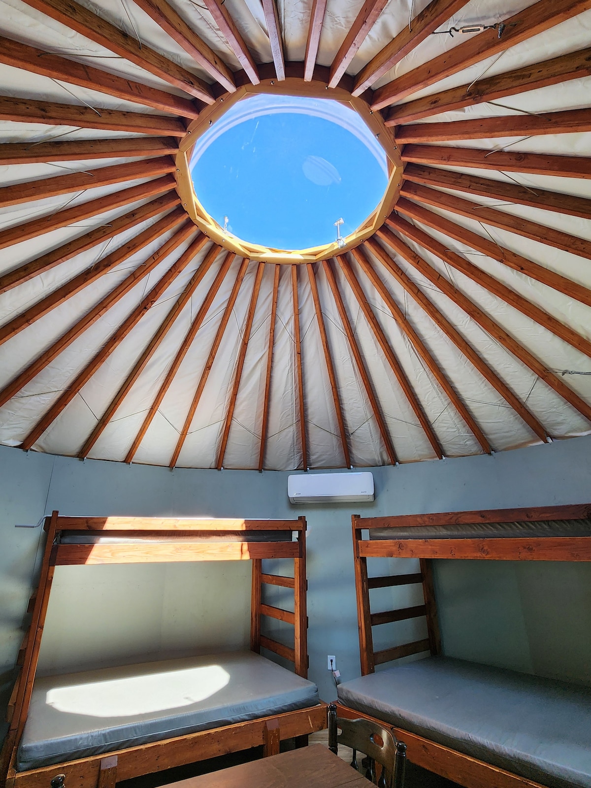 The interior of the yurt showcases a circular ceiling with wooden beams radiating from a central skylight. Two wooden bunk beds are positioned against the wall, offering padded sleeping spaces. A cooling unit is mounted for climate control, enhancing comfort within the space.