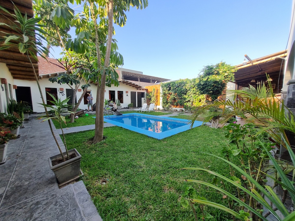 A serene outdoor area is shown, featuring a sparkling swimming pool surrounded by well-maintained grass and lush greenery. The pool is bordered by a stone path and vibrant plants. Sunlight illuminates the space, enhancing the inviting atmosphere created by the plant-filled surroundings.