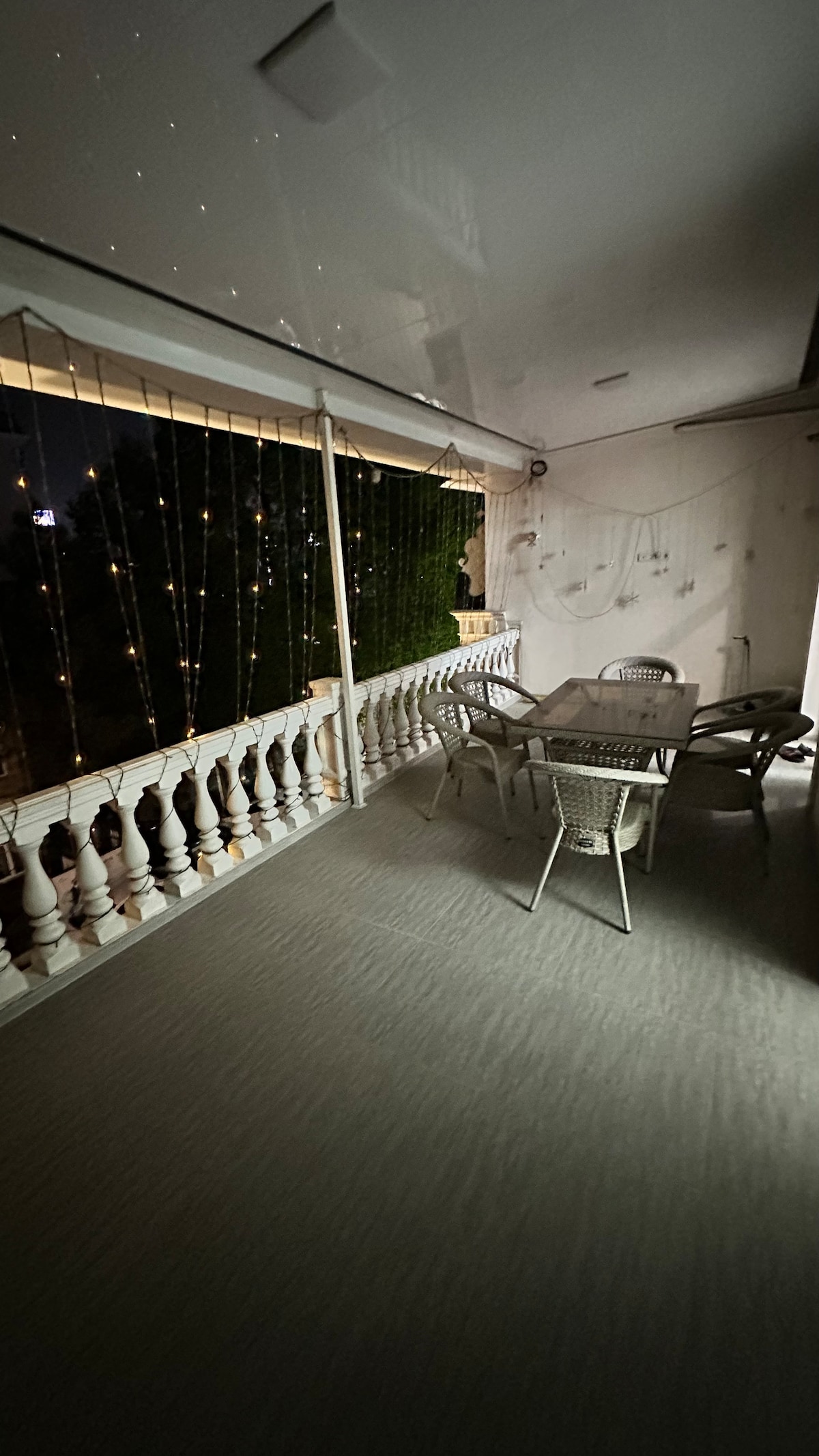 A spacious summer terrace is visible, featuring a glass table surrounded by four woven chairs. Strings of decorative lights hang above, creating a soft ambience. A balustrade borders the terrace, providing an open view of the outside while maintaining a sense of privacy.