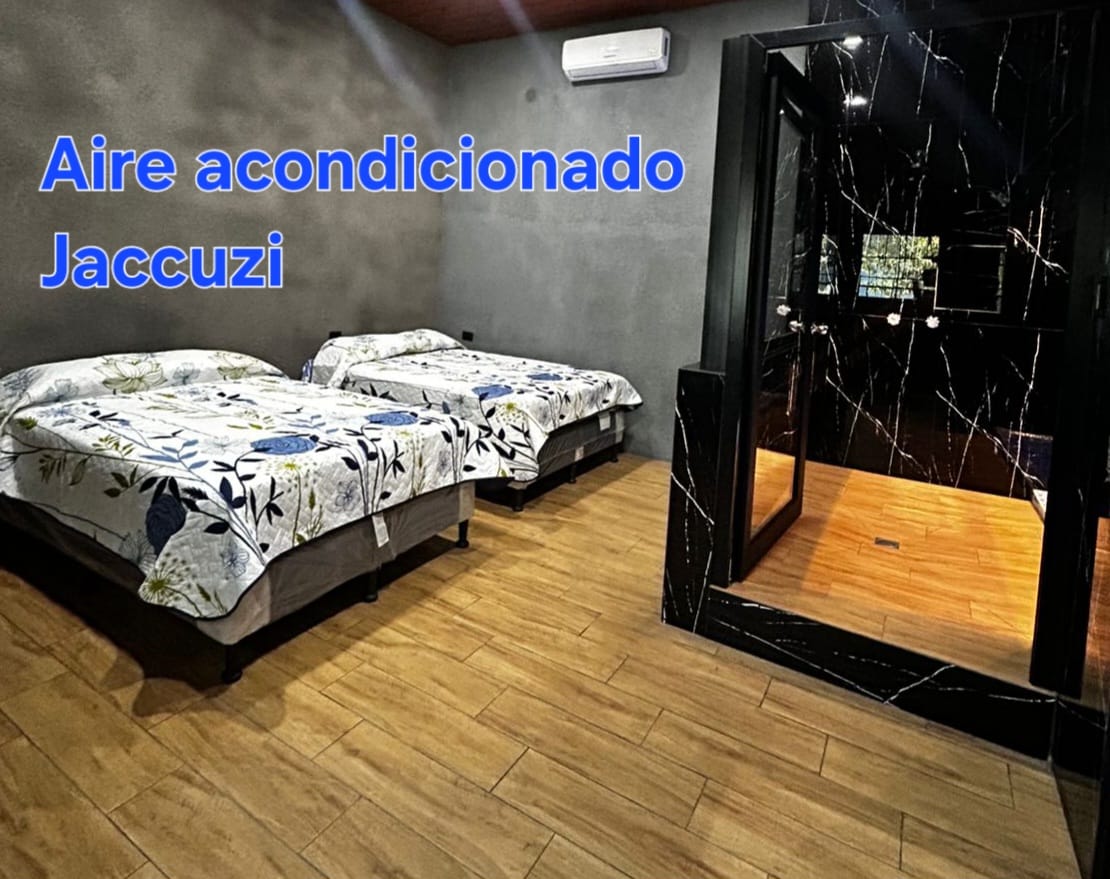 The room features two beds, each draped with floral-patterned bedding. A sleek glass shower space is visible in the corner, while wood-look flooring adds warmth. The presence of air conditioning is indicated, enhancing the comfort of the environment.