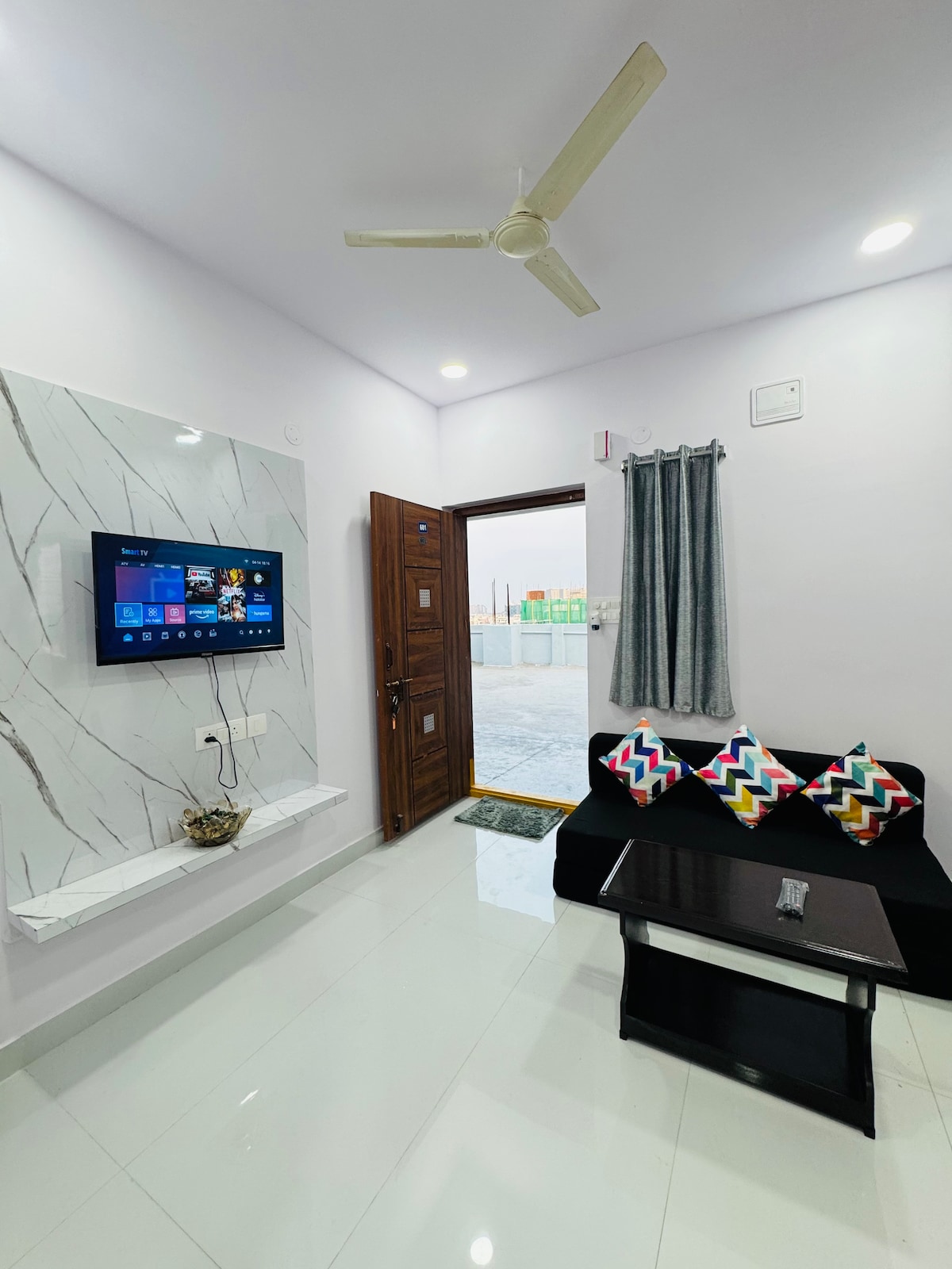 A modern living area features a black coffee table positioned in front of a sofa adorned with colorful cushions. A smart TV is mounted on a marble accent wall, while light reflects off the polished floor. A door leads to an outdoor space, framed by a gray curtain.