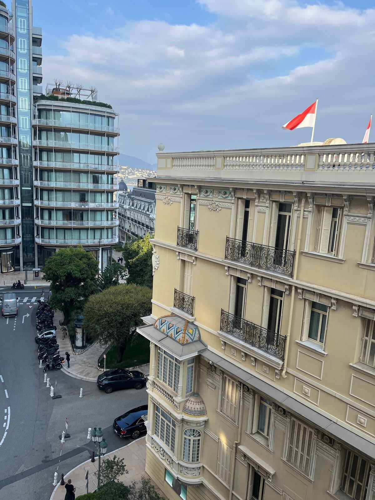 A view from the studio showcases the architectural contrast between modern high-rise buildings and classic structures. Flags flutter atop the neighboring building, while the streets below are lined with greenery and parked vehicles, indicating a vibrant urban environment.