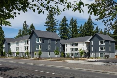 A modern multi-unit building is presented among lush greenery. The three sections feature a mix of gray and white exteriors, large windows, and well-maintained landscaping. A broad, paved area lines the front, providing easy access from the street.