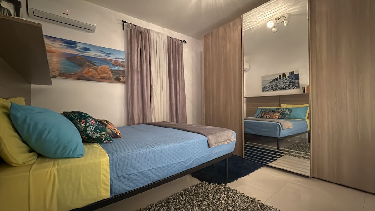 A cozy bedroom features a spacious bed with blue and yellow linens, complemented by patterned throw pillows. The room includes a large mirrored wardrobe reflecting the decor. Light-colored curtains frame a window, allowing for natural light to brighten the space.