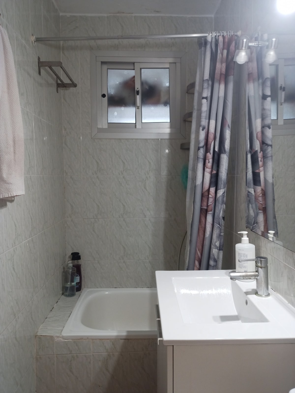 A compact bathroom features light-colored tiled walls and a small bathtub with a shower curtain displaying a soft design. A mirror above the sink reflects natural light from a window, which has two small panes. Basic toiletries are arranged on the sink's countertop.