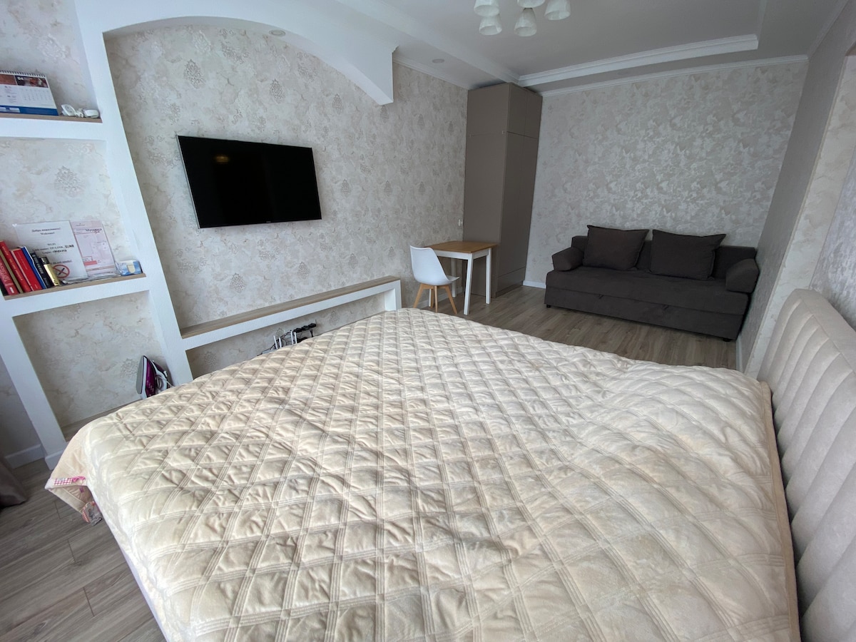 A spacious sleeping area features a large, neatly made bed with a quilted coverlet. A television is mounted on the wall opposite the bed, and a simple wooden dining table with a chair is visible nearby. A soft couch is situated against the wall, enhancing the cozy atmosphere.