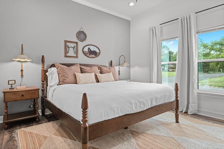 Udobna spavaća soba na farmi s bračnim krevetom s baldahinom, toplo uređenje u neutralnim bojama, nježno osvjetljenje i veliki prozori s puno prirodnog svjetla. 