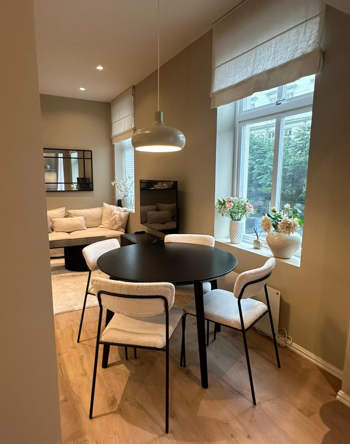 A dining area is visible, featuring a round black table surrounded by four light-colored chairs. Natural light is allowed in through a large window adorned with sheer white curtains, highlighting a cozy seating area in the background with neutral-toned furnishings and decorative plants.