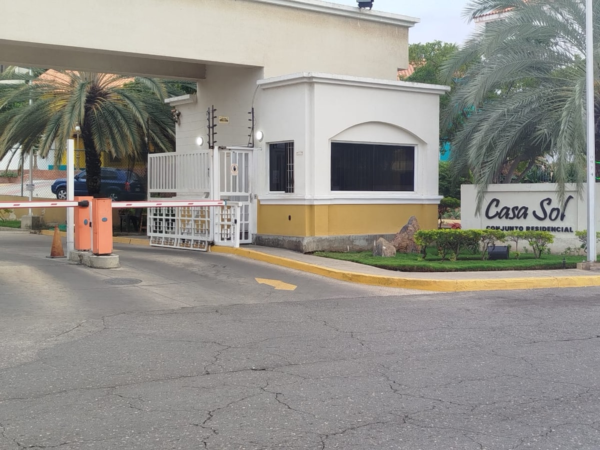 The entrance to Casa Sol is featured, showcasing a secure gate and guardhouse. Well-maintained palm trees are positioned on either side of the driveway, providing a welcoming atmosphere. A clean, paved road leads into the residential complex, enhancing accessibility.