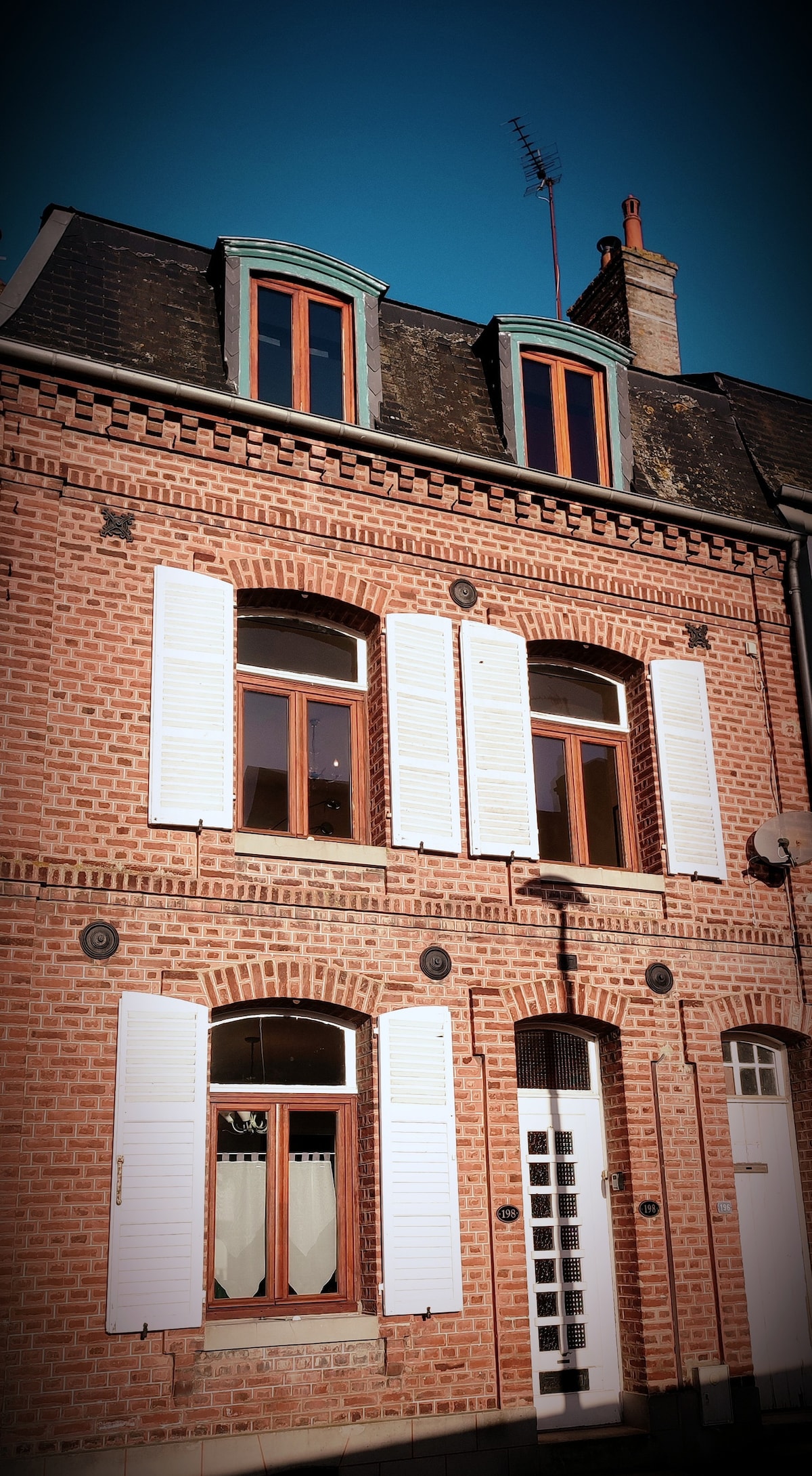 A charming brick facade features multiple windows with white shutters, creating a balanced and inviting appearance. The roofline includes classic dormer windows, adding character to the structure. A simple front door is prominently displayed, contributing to the home's welcoming entrance.
