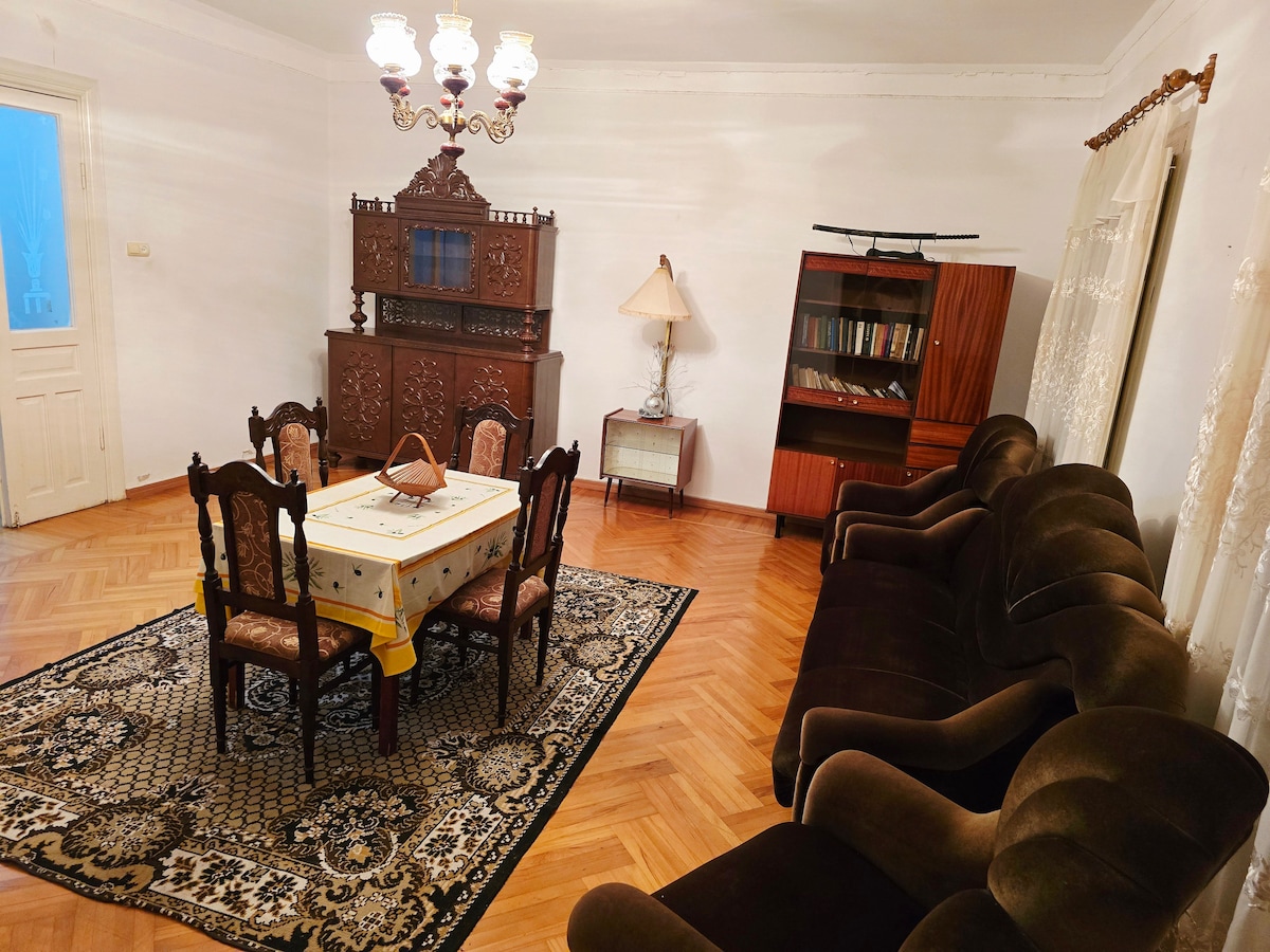 A spacious living room features dark wood furniture, including a carved cabinet and a bookshelf. A round dining table is surrounded by ornate chairs, and a plush dark green sofa is positioned near a large area rug. Natural light enters through a nearby window.