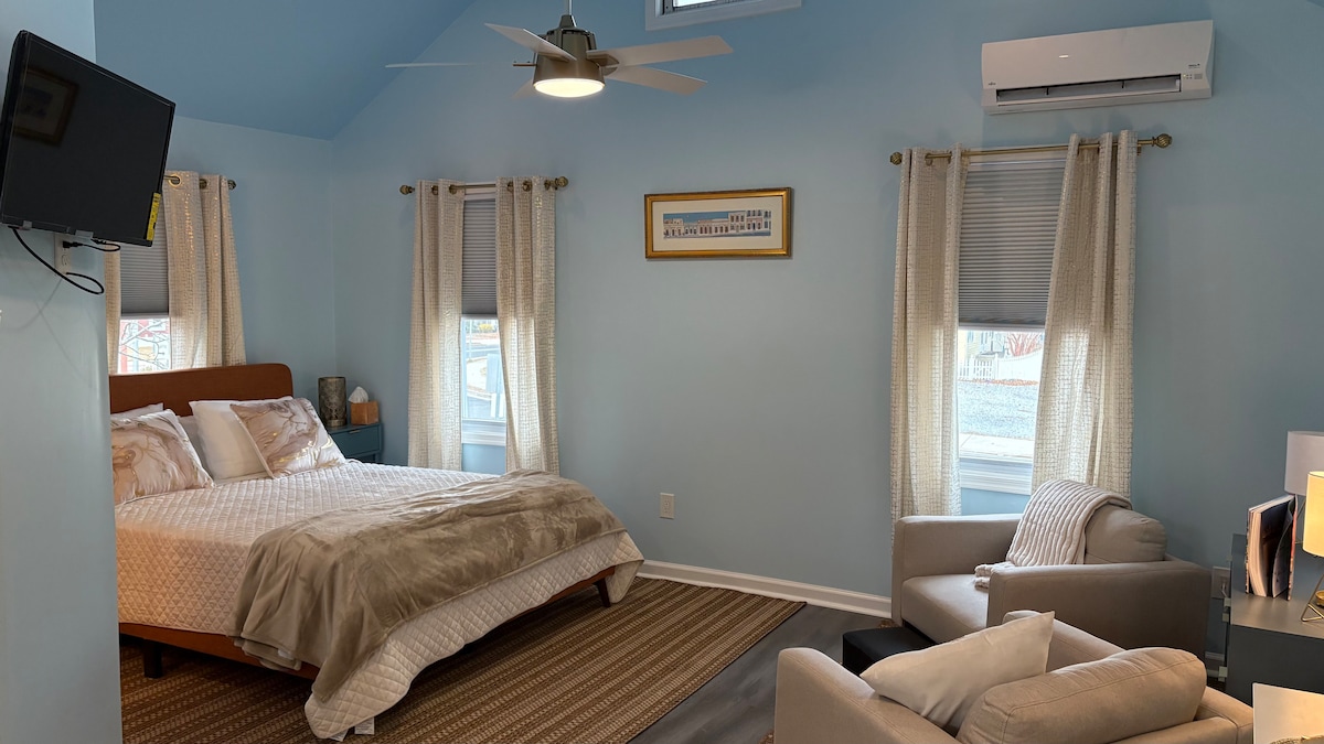 A serene sleeping area features a comfortable queen bed, accentuated by soft bedding. Natural light filters through two windows, complemented by light curtains. A cozy seating area includes an armchair, while a ceiling fan provides airflow. A wall-mounted TV is positioned for easy viewing.