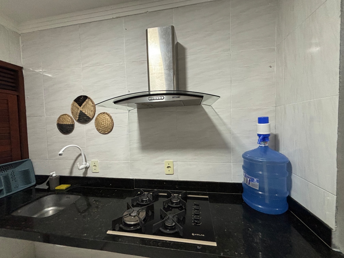A modern kitchen area is presented with a sleek black countertop and a gas stove featuring four burners. Above, a polished stainless steel hood is mounted. A water dispenser sits on the right, while decorative wall elements add visual interest to the space.