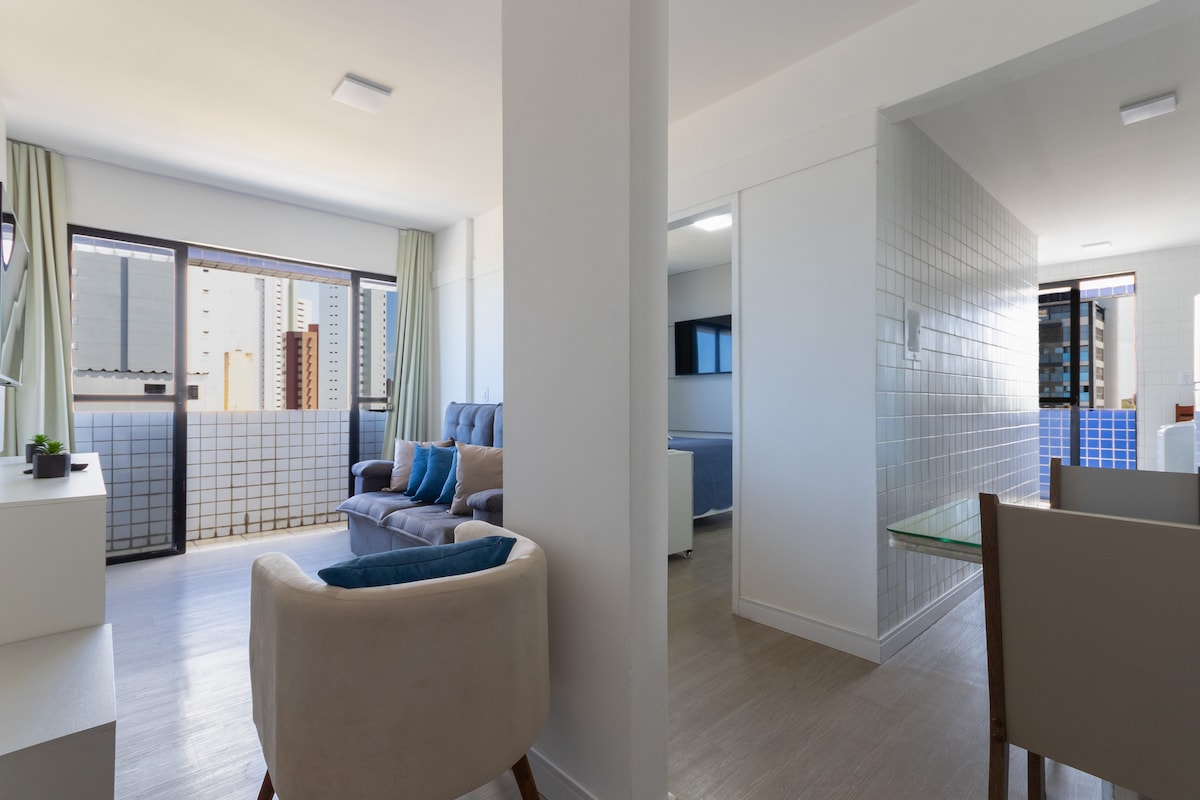 A spacious living area is featured, showcasing a comfortable seating arrangement with a light-colored armchair and a blue-accented sofa. Large windows let in natural light, offering a view of a cityscape. The tiled wall and adjacent dining area create an open and airy feel.