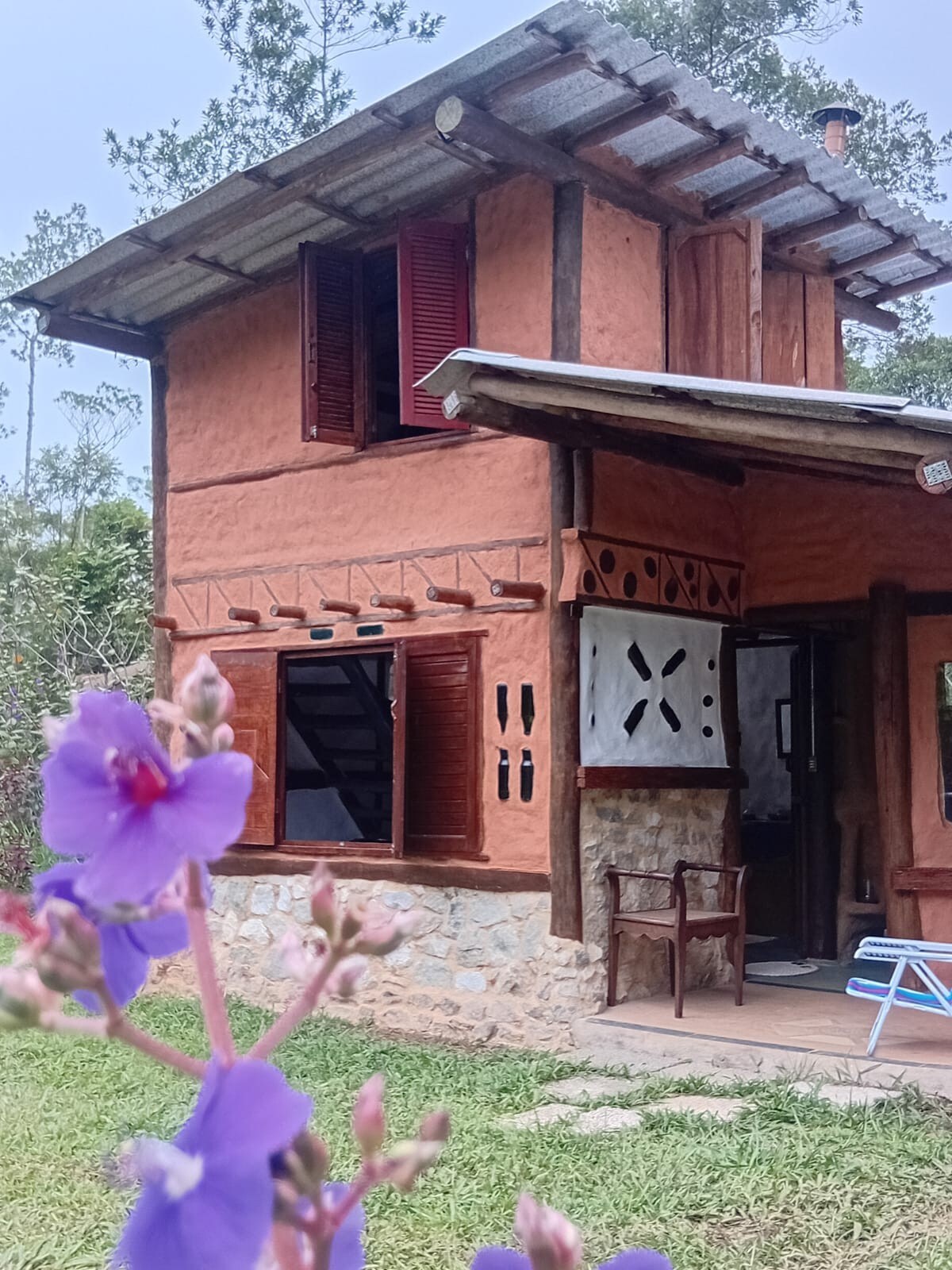 The two-story building features a warm, earthy exterior with a blend of stone and mud walls. Large, wooden shutters frame the windows, enhancing the structure. A porch area is visible with a simple chair, surrounded by vibrant purple flowers and natural greenery.