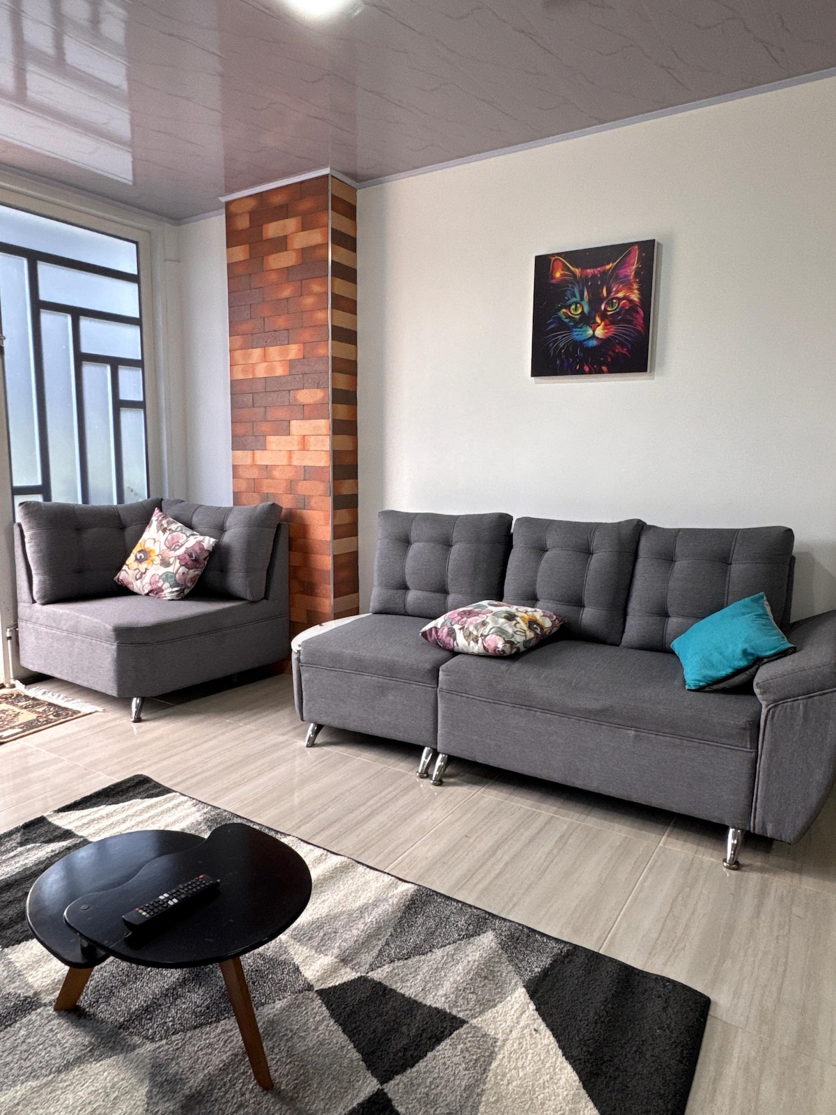 A modern living area features a gray sofa and a matching armchair, adorned with patterned cushions. A small round table rests on a textured rug, while light enters through large windows, highlighting the warm tones of a brick accent wall and a colorful cat painting.