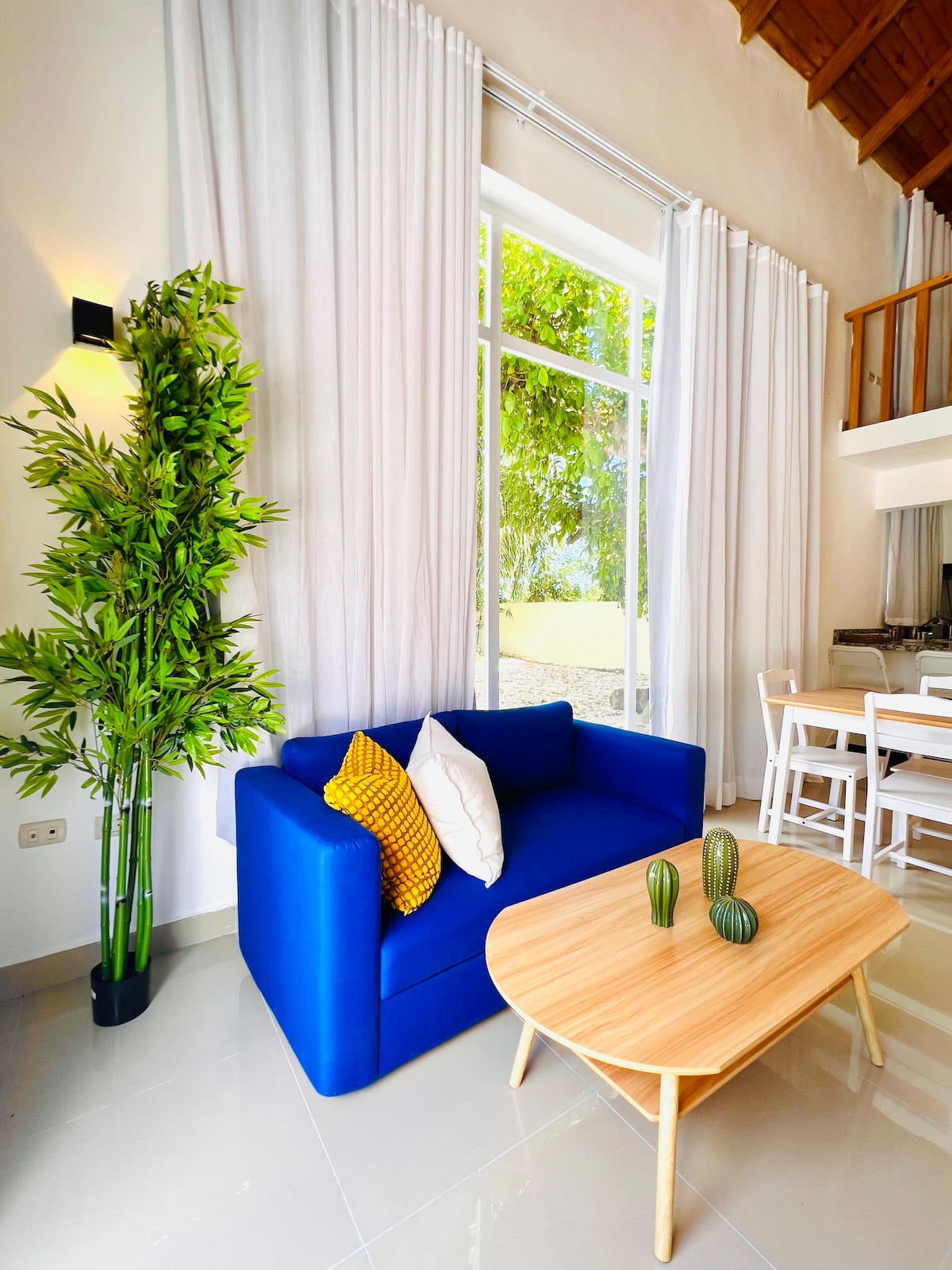 A bright seating area is presented with a blue sofa, adorned with decorative pillows. A light wooden coffee table rests in front, complemented by small plants. Tall windows allow natural light to flow in, revealing a view of the outdoors, where greenery is visible.