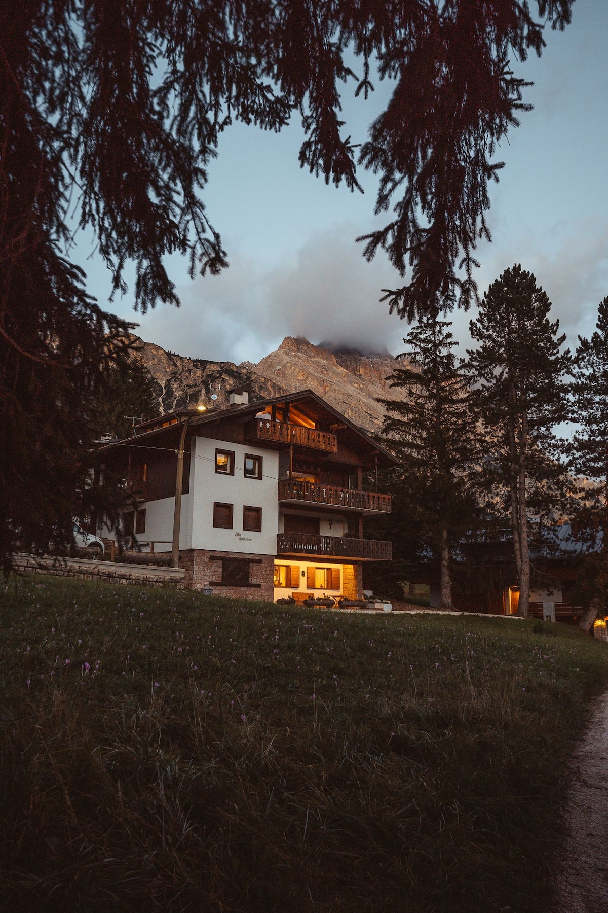 A three-story villa is set against a mountainous backdrop, highlighted by soft evening light. Large windows illuminate the interior, while a spacious lawn surrounds the property. Tall trees frame the view, adding to the serene atmosphere of the setting.