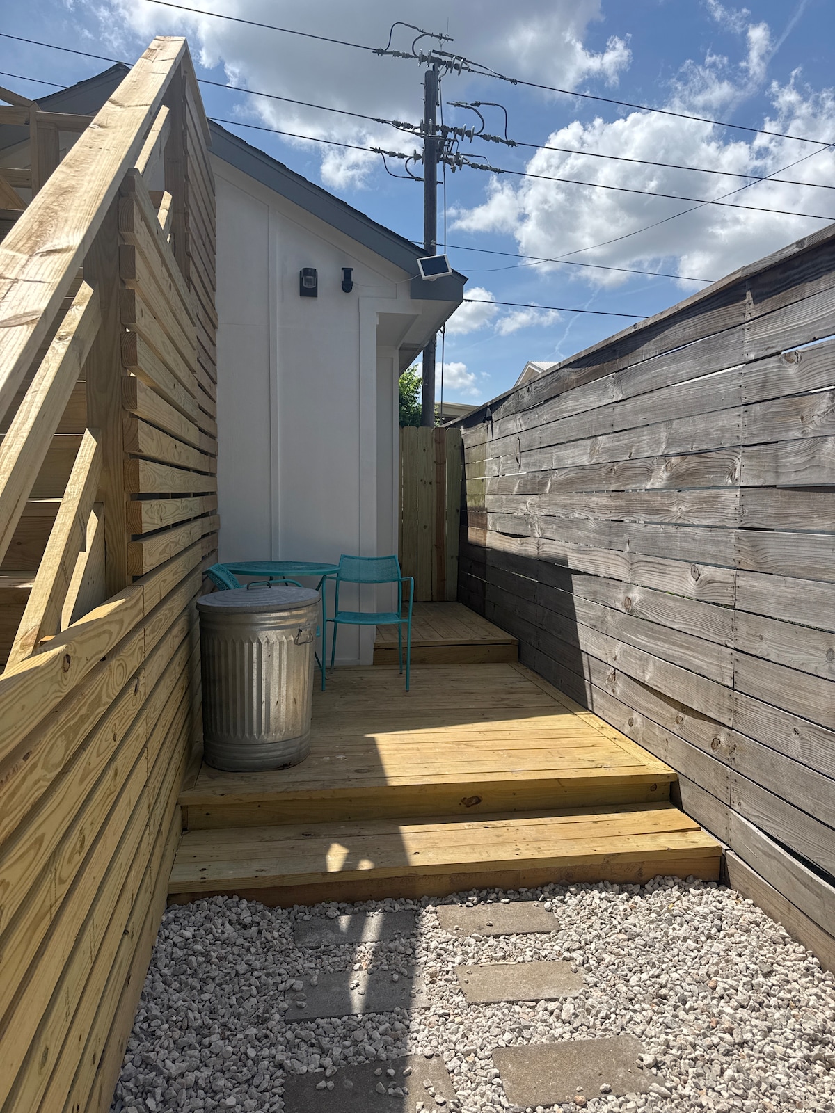 A wooden deck leads up to a private entrance, flanked by dark wooden fencing on both sides. Two turquoise chairs are positioned on the deck, with a silver trash can standing nearby. The area is illuminated by natural light, with scattered clouds visible above.