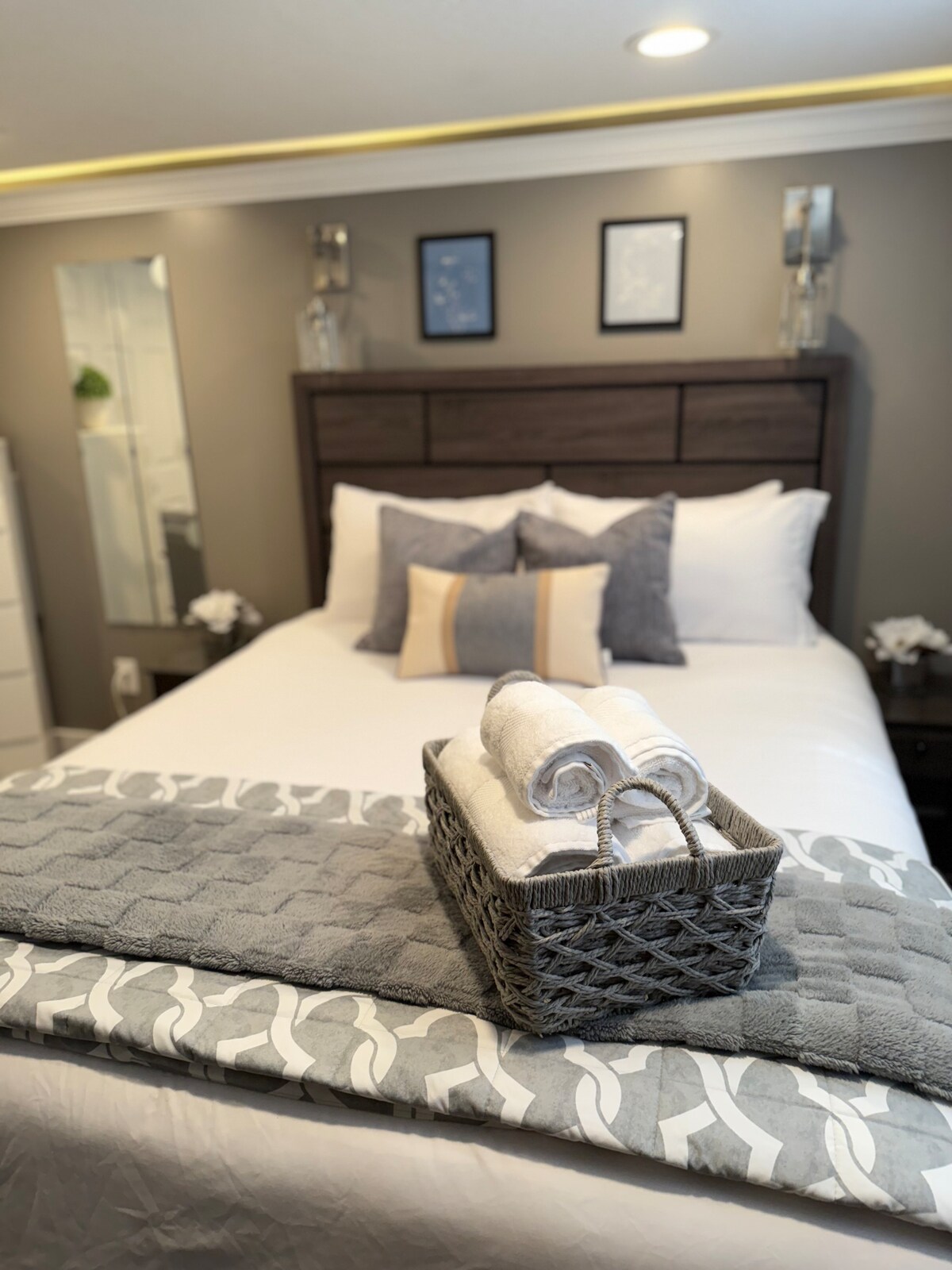 A neatly arranged sleeping area is highlighted by a spacious bed dressed in crisp white linens. A decorative basket holds rolled towels, sitting atop a textured blanket. Framed artwork and a large mirror adorn the muted gray walls, complemented by soft lighting.