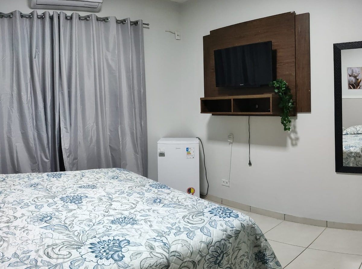 A private bedroom is illustrated, featuring a spacious bed with a patterned coverlet. A wall-mounted television is positioned above a small shelf, while a mini-fridge is discreetly placed to one side. Light grey curtains frame the window, and a decorative mirror adds depth to the space.